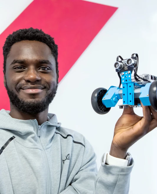 Student in a grey hoodie holding up a blue robotic vehicle, smiling against a colourful geometric wall. Student in a grey hoodie holding up a blue robotic vehicle, smiling against a colourful geometric wall.